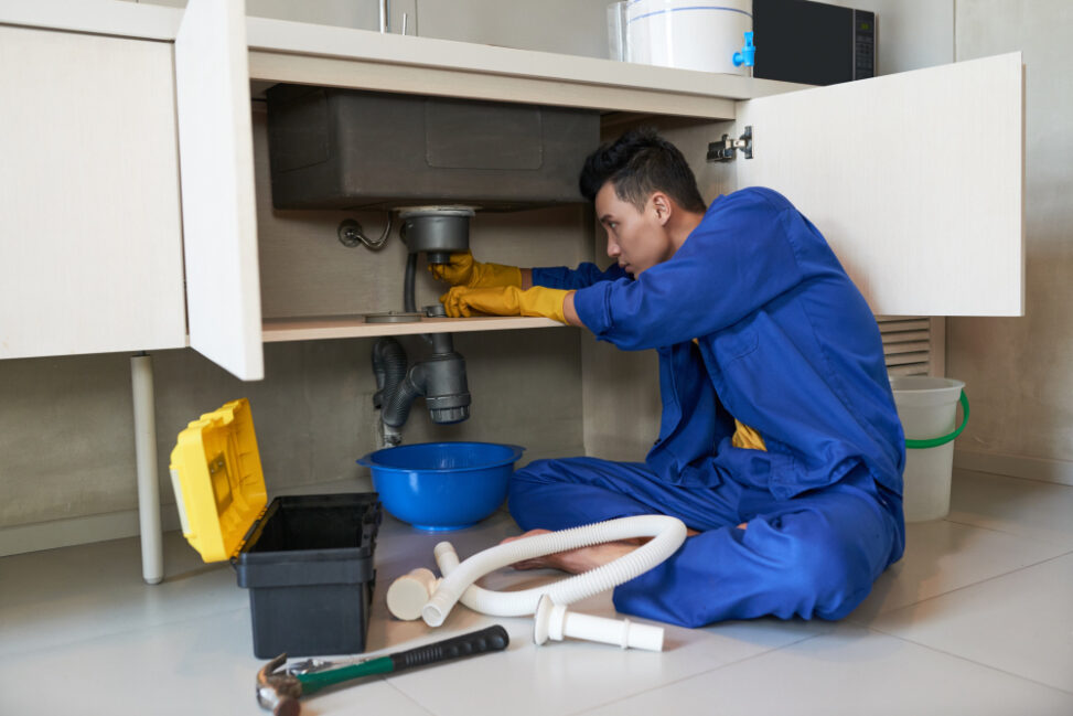asian-plumber-blue-overalls-clearing-blockage-drain-973x649